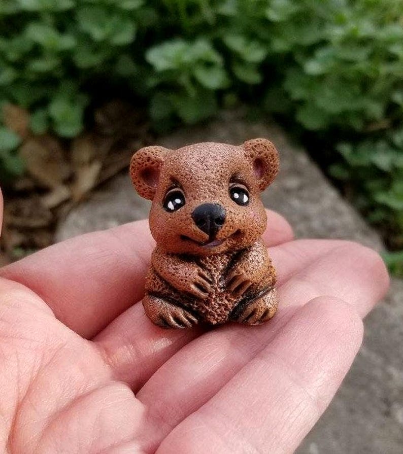 May include: A small, textured brown figurine of a quokka with rounded ears, large dark eyes, and a black nose. The creature is depicted in a seated position with its front paws resting together near its chest, featuring a smooth, matte finish.