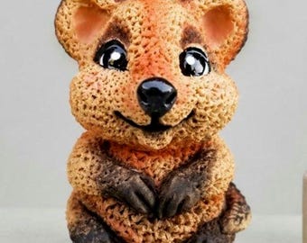 May include: A small, textured figurine of a quokka with a light brown, porous surface. The creature features large, dark, glossy eyes, a prominent black nose, and a subtle smile. Its paws and feet are dark brown, contrasting with its lighter body.