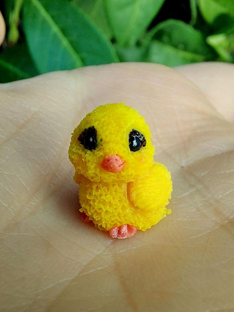 May include: A small, yellow chick figurine with black eyes and an orange beak and feet. The chick has a textured, fluffy appearance, and is likely a decorative item or toy.