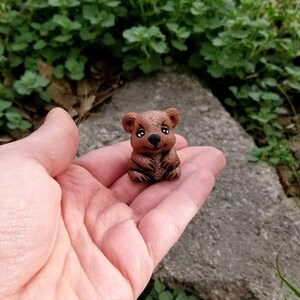 May include: A small, brown, sculpted figurine of a quokka with a rounded head, pointed ears, a black nose, and large, dark eyes. The creature is depicted in a seated position with its front paws resting against its chest, showing a textured surface.