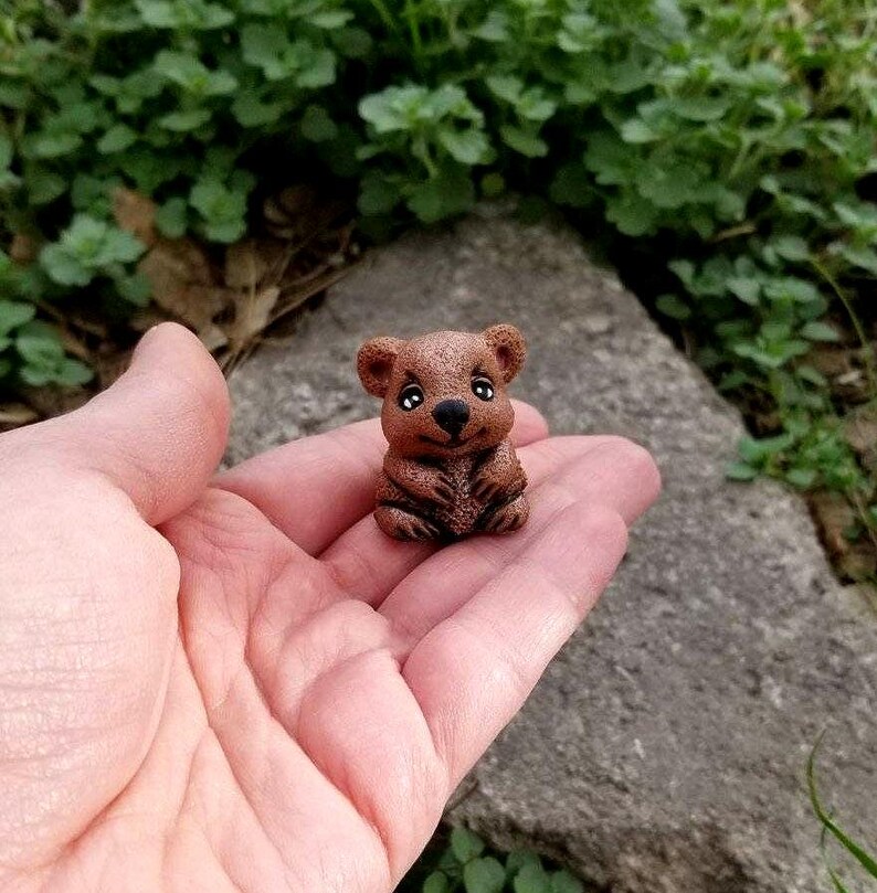 May include: A small, brown, sculpted figurine of a quokka with a rounded head, pointed ears, a black nose, and large, dark eyes. The creature is depicted in a seated position with its front paws resting against its chest, showing a textured surface.