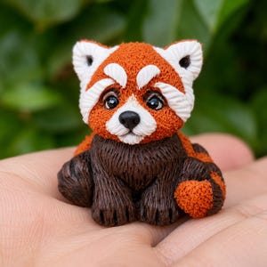 Peut inclure: Une petite figurine de panda roux sculptée avec une tête et un dos texturés orange rouille, des marques faciales blanches, des yeux noirs et une poitrine ainsi que des membres brun foncé. La figurine présente un aspect détaillé et poilu.