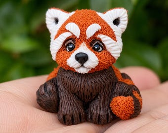 May include: A small, sculpted red panda figurine featuring a textured rust-orange head and back with white facial markings, black eyes, and a dark brown chest and limbs. The figure has a detailed, fur-like appearance with a curled, striped tail.