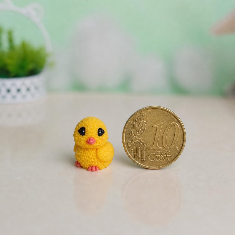 May include: A small, yellow chick figurine with black eyes and a pink beak and feet. A gold-colored 10 Euro cent coin is next to the chick. The chick is made of a textured material.