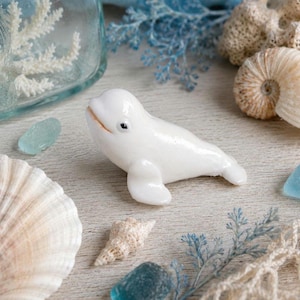 May include: A glossy white ceramic beluga whale ornament, positioned amongst seashells, blue sea glass, and coral. The scene is set on a light wooden surface, creating a coastal aesthetic.