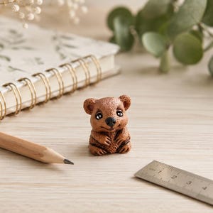 May include: A small, brown, textured figurine of a quokka with large black eyes and a smiling expression. The figurine is positioned next to a wooden pencil and a metal ruler that shows a measurement of approximately 3 centimetres in length.