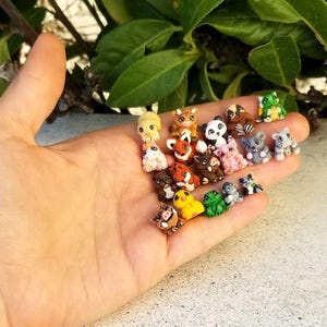 May include: A collection of miniature animal figurines, including a panda, fox, and pig, in various colors. The figurines are small, detailed, and appear to be handmade. The collection is held in a hand, with green foliage in the background.