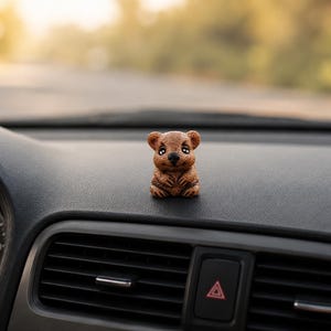 May include: A small, brown, textured figurine of a quokka with large dark eyes and a smiling expression, positioned on the dashboard of a vehicle near the air vents and the speedometer gauge.