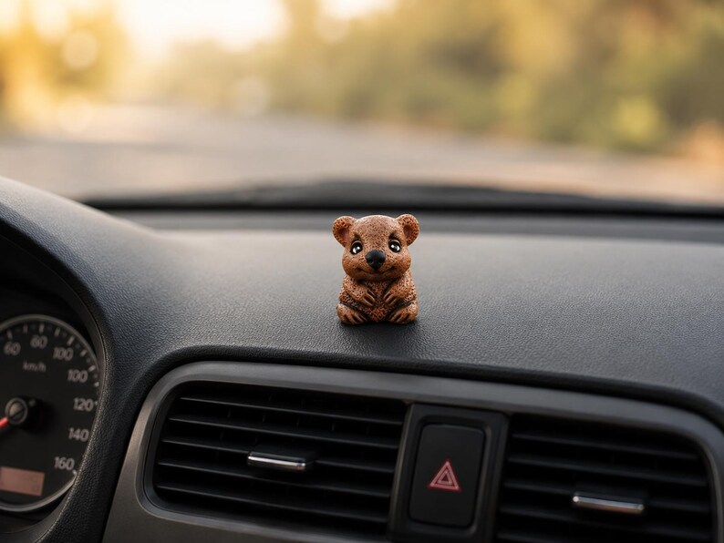 May include: A small, brown, textured figurine of a quokka with large dark eyes and a smiling expression, positioned on the dashboard of a vehicle near the air vents and the speedometer gauge.
