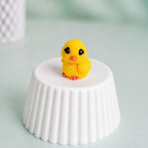 May include: A small, yellow chick figurine with black eyes and an orange beak and feet, perched on a white, fluted, ceramic pedestal. The chick is a detailed, handcrafted item, likely for decorative purposes.