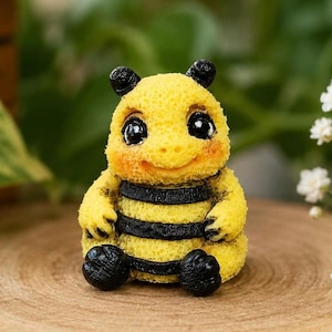 Puede incluir: Una pequeña figura de abeja amarilla texturizada con grandes ojos negros, una boca sonriente y dos rayas horizontales negras en el cuerpo. La abeja tiene antenas, manos y pies negros, con una textura de superficie porosa en todo el cuerpo.