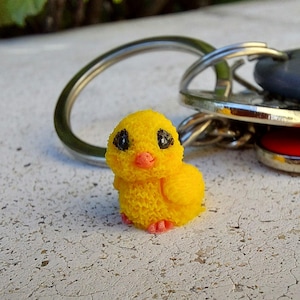 May include: A yellow chick keychain with black eyes and a pink beak and feet. The chick is attached to a silver key ring with other keys. The chick is made of a textured material.