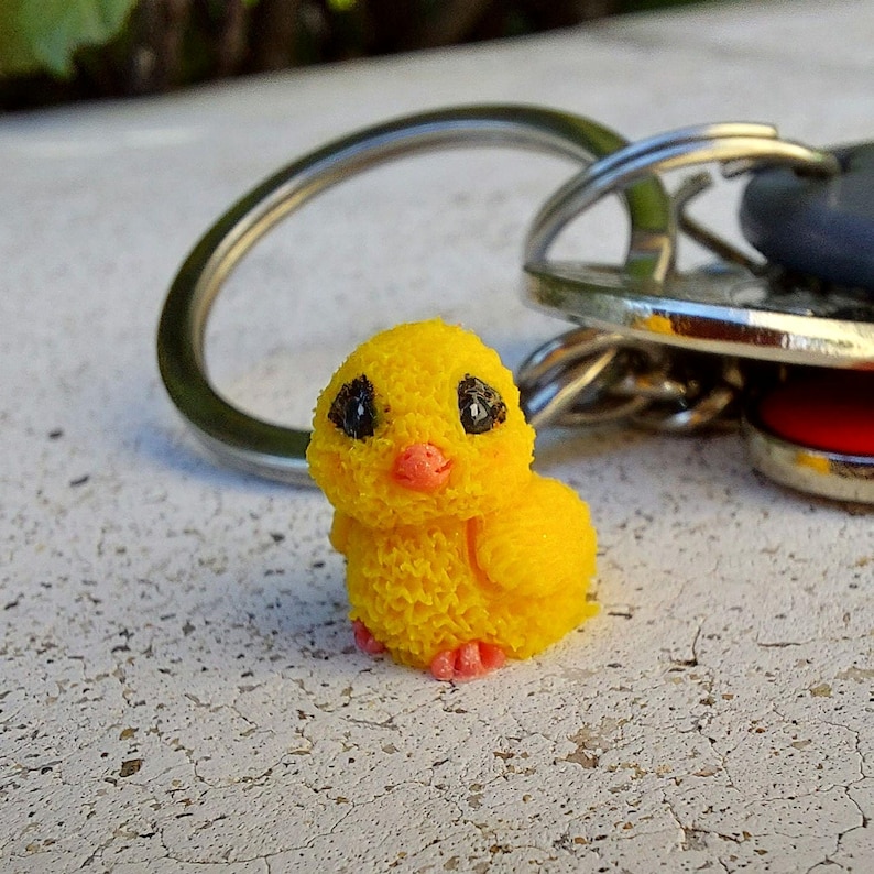 May include: A yellow chick keychain with black eyes and a pink beak and feet. The chick is attached to a silver key ring with other keys. The chick is made of a textured material.