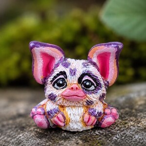 May include: A small, hand-painted creature figurine with large, pointed ears, a white base coat, and purple and orange mottled spots. It features large black eyes, a pink nose, and pink paws, with a textured, fur-like appearance throughout.