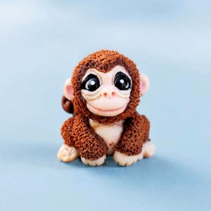 Puede incluir: Pequeña figura de mono marrón con ojos grandes y expresivos y una expresión sonriente. El mono tiene un aspecto de pelaje texturizado y está sentado sobre sus cuartos traseros. El fondo es azul claro liso.