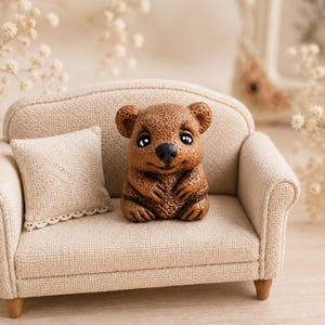May include: A miniature brown bear figurine with a textured surface, dark eyes, and a black nose, sitting on a small beige fabric sofa. The sofa features a matching square throw pillow with a delicate lace trim along the bottom edge.