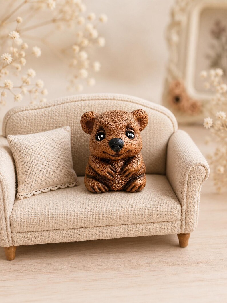 May include: A miniature brown bear figurine with a textured surface, dark eyes, and a black nose, sitting on a small beige fabric sofa. The sofa features a matching square throw pillow with a delicate lace trim along the bottom edge.
