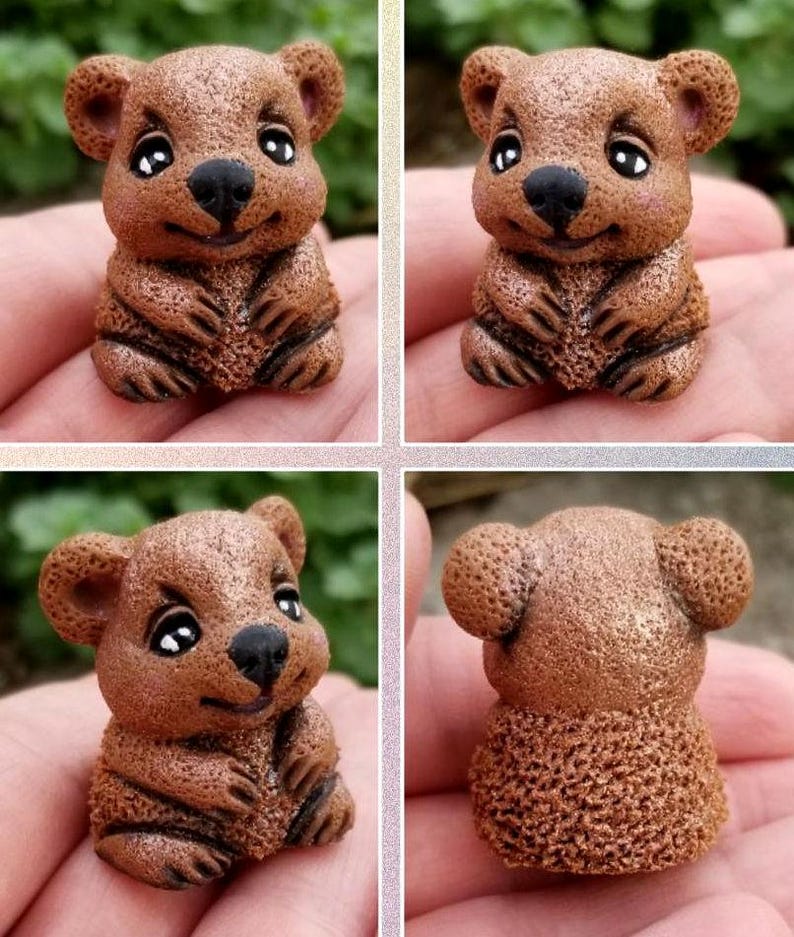 May include: A small, brown, textured figurine of a quokka with large, dark, glossy eyes, a black nose, and a smiling expression. The figure has rounded ears and a detailed, porous surface texture, shown from multiple angles in a four-part grid.