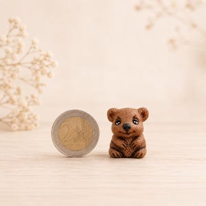 May include: A small, brown, textured figurine of a koala with large ears, dark eyes, and a black nose, positioned next to a 2 euro coin for scale. The koala is sitting in an upright position with its front paws resting on its chest.