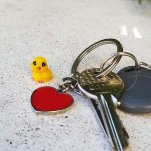 May include: A keychain with a yellow chick charm, a red heart charm, and keys. The chick charm is small and detailed, with black eyes and a textured surface. The heart charm is red with a silver border. The keys are attached to a silver ring.