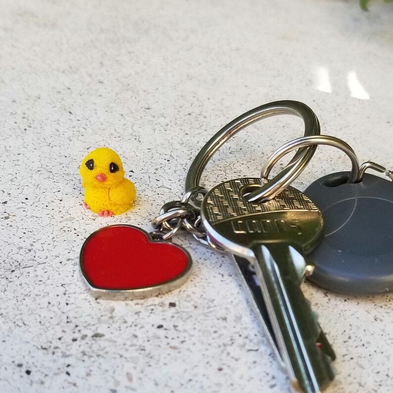 May include: A keychain with a yellow chick charm, a red heart charm, and keys. The chick charm is small and detailed, with black eyes and a textured surface. The heart charm is red with a silver border. The keys are attached to a silver ring.