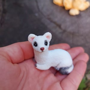 May include: A small white figurine of a weasel with black eyes and a black nose. The figurine is sitting on a person's hand.