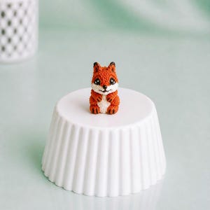 Könnte beinhalten: Eine kleine orangefarbene Fuchsskulptur mit schwarzen Akzenten und weißen Details sitzt auf einem weißen, geriffelten Sockel. Der Fuchs hat einen lächelnden Gesichtsausdruck. Der Hintergrund ist ein sanftes, gedämpftes Grün. Der Sockel hat etwa 10 cm Durchmesser.
