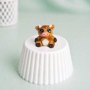 May include: A small, handmade cow figurine with brown and white fur, tiny horns, and large eyes. The cow is placed on a white, fluted ceramic pedestal. The background is a soft, muted green.
