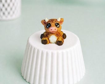 May include: A miniature, handcrafted cow figurine with brown and white fur, small horns, and large, expressive eyes. The cow sits atop a white, fluted, ceramic pedestal. The background is a soft, muted green.