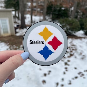 May include: A round sticker with a gray border and a white background. The sticker features the Pittsburgh Steelers logo, which includes a yellow, red, and blue star design. The word "Steelers" is printed in black text below the star design.