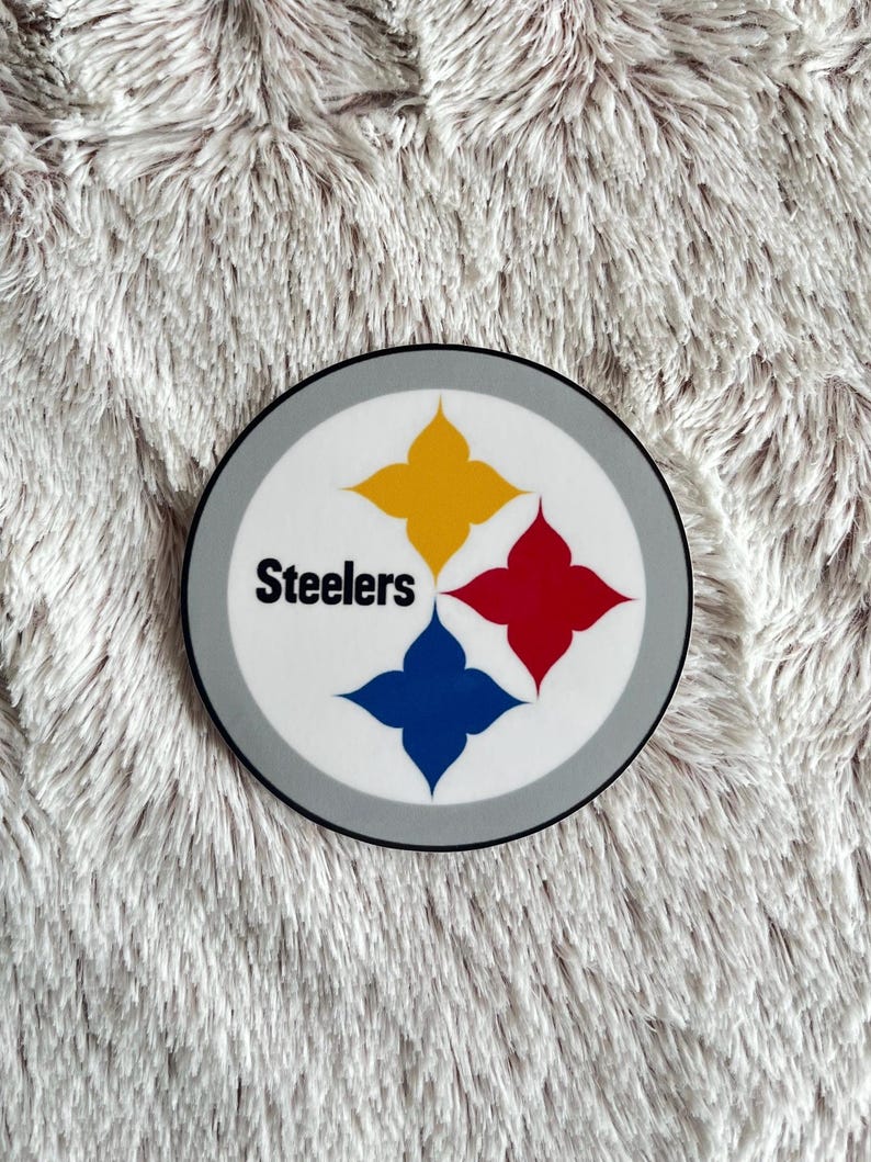 May include: A white and gray circular sticker with the Pittsburgh Steelers logo. The logo features a black star with four points, each point is a different color: yellow, red, blue, and yellow. The word "Steelers" is written in black text above the star.
