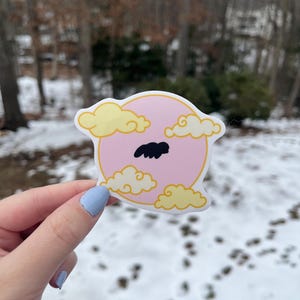 May include: A pink and white sticker with a yellow outline. The sticker features a cartoon sun with clouds and a black mustache.