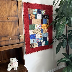 May include: A red and white patchwork quilt with a variety of colorful patterns hangs on a white wall. The quilt is made up of squares of different fabrics, each with a unique design. The quilt is framed by a red border.