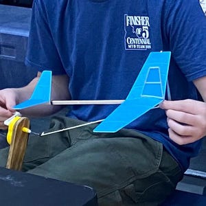 May include: A blue and white model airplane is held by a person. The plane has a blue body and wings, with a white support beam. The model is held over a wooden and yellow base. The person is wearing a blue shirt with text.