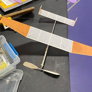 Pode incluir: Dois modelos de avião com asas laranja e brancas são exibidos. Um é maior, com uma envergadura de aproximadamente 46 cm, e o outro é menor. Uma hélice de madeira e um recipiente de plástico transparente com materiais de artesanato também são visíveis.