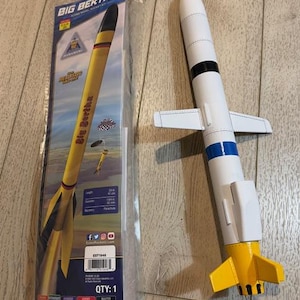 May include: A Big Bertha model rocket kit, with a yellow and black rocket in its packaging and a fully assembled white, blue, and yellow rocket. The packaging displays the rocket's name and a graphic of the rocket in flight. The assembled rocket has fins and a nose cone.