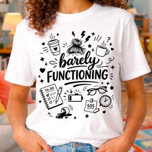 Puede incluir: Camiseta blanca con un diseño gráfico negro. El diseño incluye una taza de café, un moño desordenado, una lista de tareas, gafas, un reloj y las palabras "barely FUNCTIONING". Una prenda casual.