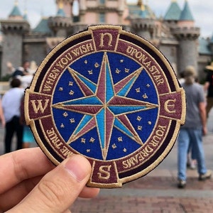 May include: A round, embroidered patch with a compass design. The compass has a blue background with a gold star in the center. The text around the compass reads "When you wish upon a star, make true your dreams".