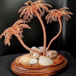 Puede incluir: Escultura de alambre de cobre de tres palmeras con frondas detalladas. Los árboles están montados sobre una base de madera con conchas marinas y arena, creando una escena de playa en miniatura. La escultura se encuentra sobre un fondo oscuro.