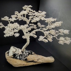 May include: A silver wire bonsai tree sculpture. The tree has a twisted trunk and delicate, detailed branches with leaves. The sculpture is mounted on a piece of driftwood with a white and black stone accent. The background is black.