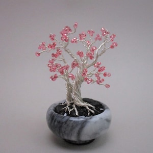 May include: A wire sculpture of a bonsai tree with pink beads for blossoms, sitting in a gray and white marble pot.