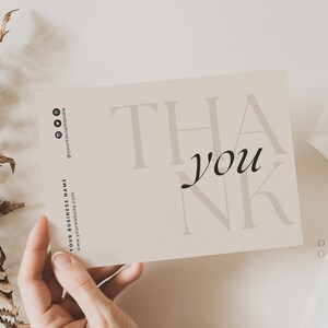 Op de afbeelding: Een eenvoudige bedankkaart met het woord "dankjewel" in een modern lettertype. De kaart is beige met zwarte tekst en wordt in een hand gehouden.