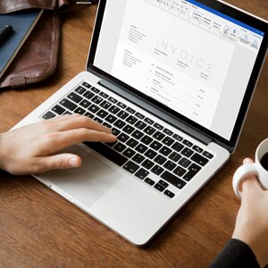 May include: A person working on a laptop computer with a white background. The laptop screen shows an invoice with the word "INVOICE" at the top. The person is wearing a watch and has a cup of coffee in their other hand.