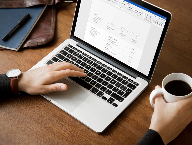 May include: A person working on a laptop computer with a white background. The laptop screen shows an invoice with the word "INVOICE" at the top. The person is wearing a watch and has a cup of coffee in their other hand.