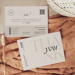 Op de afbeelding: Een witte bedankkaart met zwarte tekst die "dankjewel" zegt. De kaart heeft een QR-code en social media-handvatten erop gedrukt. De kaart ligt op een bruine stoffen ondergrond.