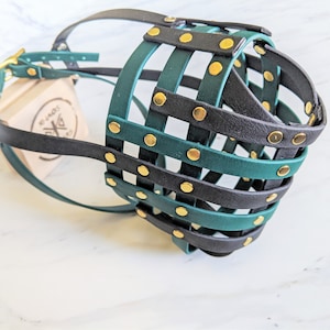 May include: A basket-style dog muzzle constructed from intersecting black and dark green leather straps secured with gold-toned metal rivets. The muzzle features an adjustable strap with a gold-toned buckle and is positioned near a small wooden block.