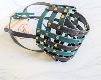 May include: A basket-style dog muzzle constructed from intersecting black and dark green leather straps secured with gold-toned metal rivets. The muzzle features an adjustable strap with a gold-toned buckle and is positioned near a small wooden block.