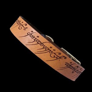 Puede incluir: Una pulsera de cuero marrón claro con escritura grabada. La pulsera presenta un cierre plateado y se encuentra sobre un fondo negro. La escritura parece estar en una fuente estilizada.