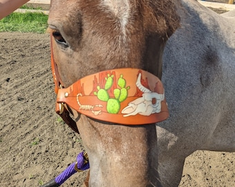 May include: A brown horse wearing a leather browband with a desert-themed design. The browband features a painted desert landscape with cacti, a skull, and a scorpion. The horse is attached to a purple lead rope.