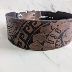 Könnte beinhalten: Breites braunes Lederhalsband für Hunde mit aufwendigen schwarzen Stammesmustern und den Buchstaben "USO". Das Halsband hat eine schwarze Innenseite und silberfarbene Metallbeschläge. Die Breite beträgt ca. 7,6 cm.